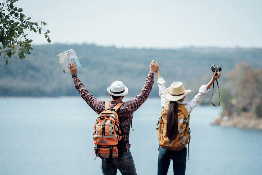 Dois viajantes de costas, um homem e uma mulher, estão de pé em frente a um lago cercado por uma paisagem verde e montanhosa. Ambos vestem chapéus e mochilas. O homem segura um mapa em uma mão enquanto a outra está levantada, e a mulher segura uma câmera em uma mão, também com o braço erguido. Eles parecem estar celebrando o momento e apreciando a vista ao ar livre.